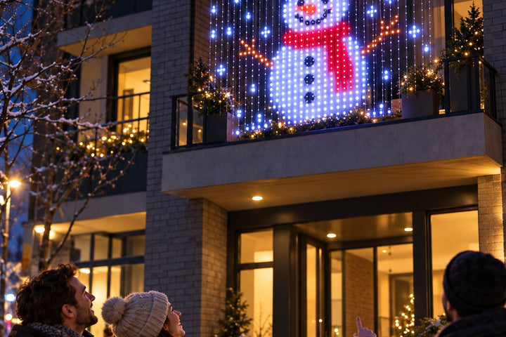 People on a snowy street pointing up at a pixel snowman on window curtain lights hung across a balcony in front of floor-to-ceiling glass